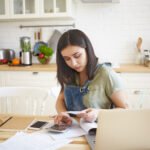 Young woman sitting in a bright kitchen, focused on debt management tasks as she reviews bills, uses a calculator, and works on her laptop, with fresh fruits and a tidy workspace in the background