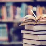 Stack of books with an open book on top in a library, perfect for mastering life skills, with a blurred background of colorful books on shelves.