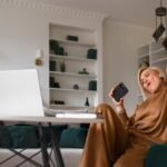 woman in comfortable brown loungewear sits in a modern home office, holding a smartphone and celebrating a success while working remotely on her laptop. 10 Popular Ways to Make Money in 2025