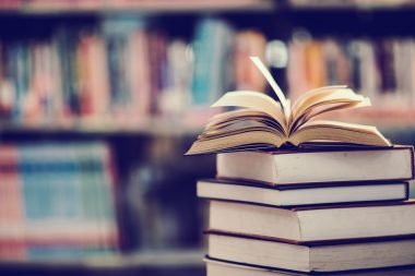 Stack of books with an open book on top in a library, perfect for mastering life skills, with a blurred background of colorful books on shelves.