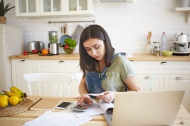 Young woman sitting in a bright kitchen, focused on debt management tasks as she reviews bills, uses a calculator, and works on her laptop, with fresh fruits and a tidy workspace in the background