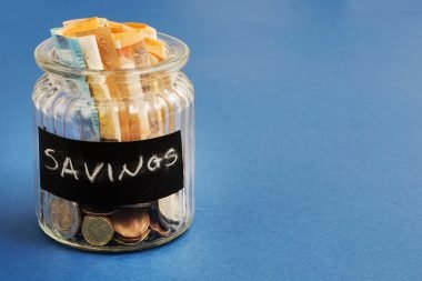 Glass jar labeled 'Savings' filled with euro banknotes and coins, symbolizing saving money and financial planning, against a blue background.