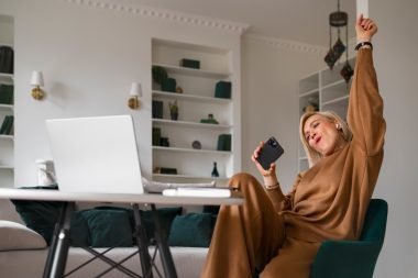 woman in comfortable brown loungewear sits in a modern home office, holding a smartphone and celebrating a success while working remotely on her laptop. 10 Popular Ways to Make Money in 2025